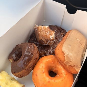 A white box holding a chocolate old fashioned, an apple fritter, a maple bar and an orange glazed donut