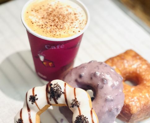 A latte beside a white glazed mochi donut, a blueberry cake donut and a croissant donut