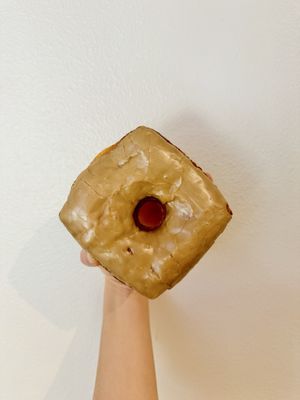 A hand holding a square maple-glazed donut with a red jelly centre against a white wall